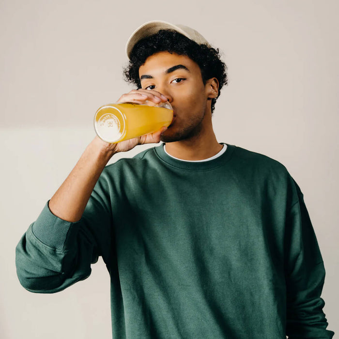 Man drinking from a glass bottle with a beige cap on a plain background.