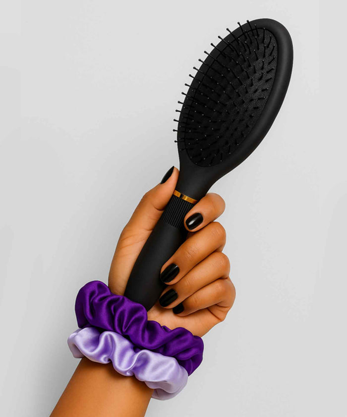 A hand with black nail polish holds a black hairbrush, wrist adorned with two pastel scrunchies—one purple, one lavender—against a plain light background.