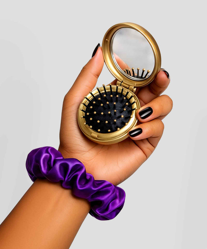 A hand with black nail polish and a purple satin scrunchie holds a gold compact with a mirror and round black hairbrush with gold bristles.