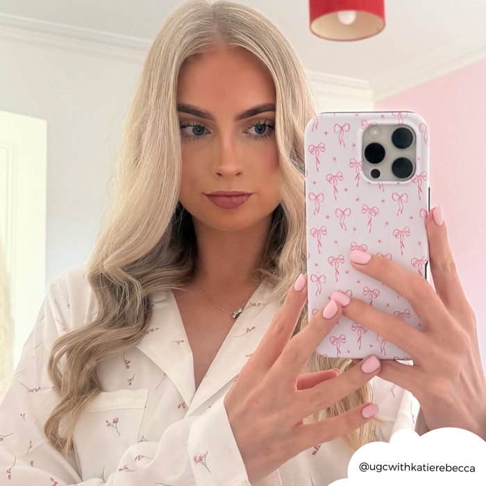 Woman with long blonde hair takes a mirror selfie holding a Coconut Lane tough phone case with a dainty bows pattern.